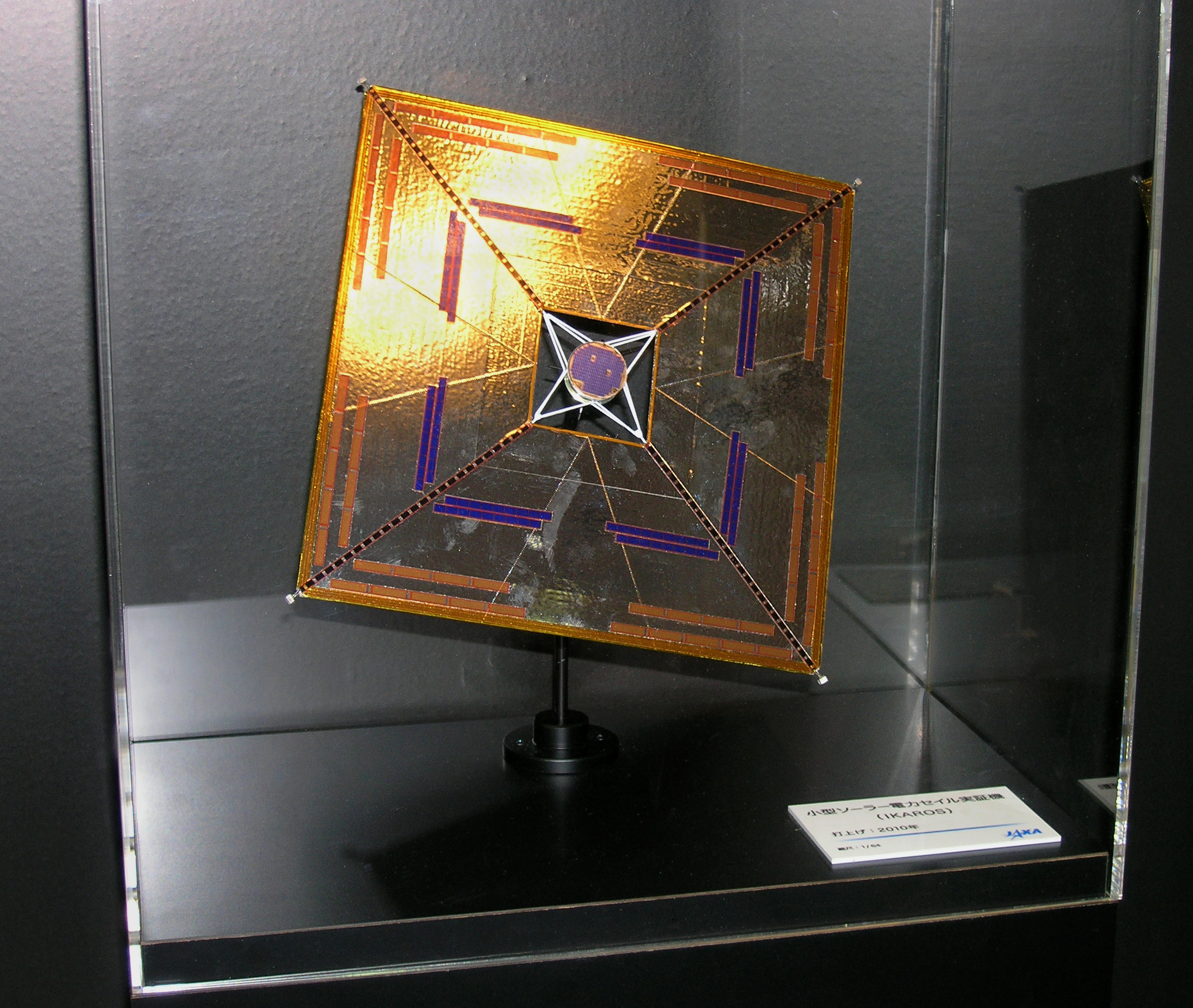 Figure shows a 1:64 scale model of the IKAROS spacecraft, a square-shape resembling a golden kite, displayed inside a glass case.